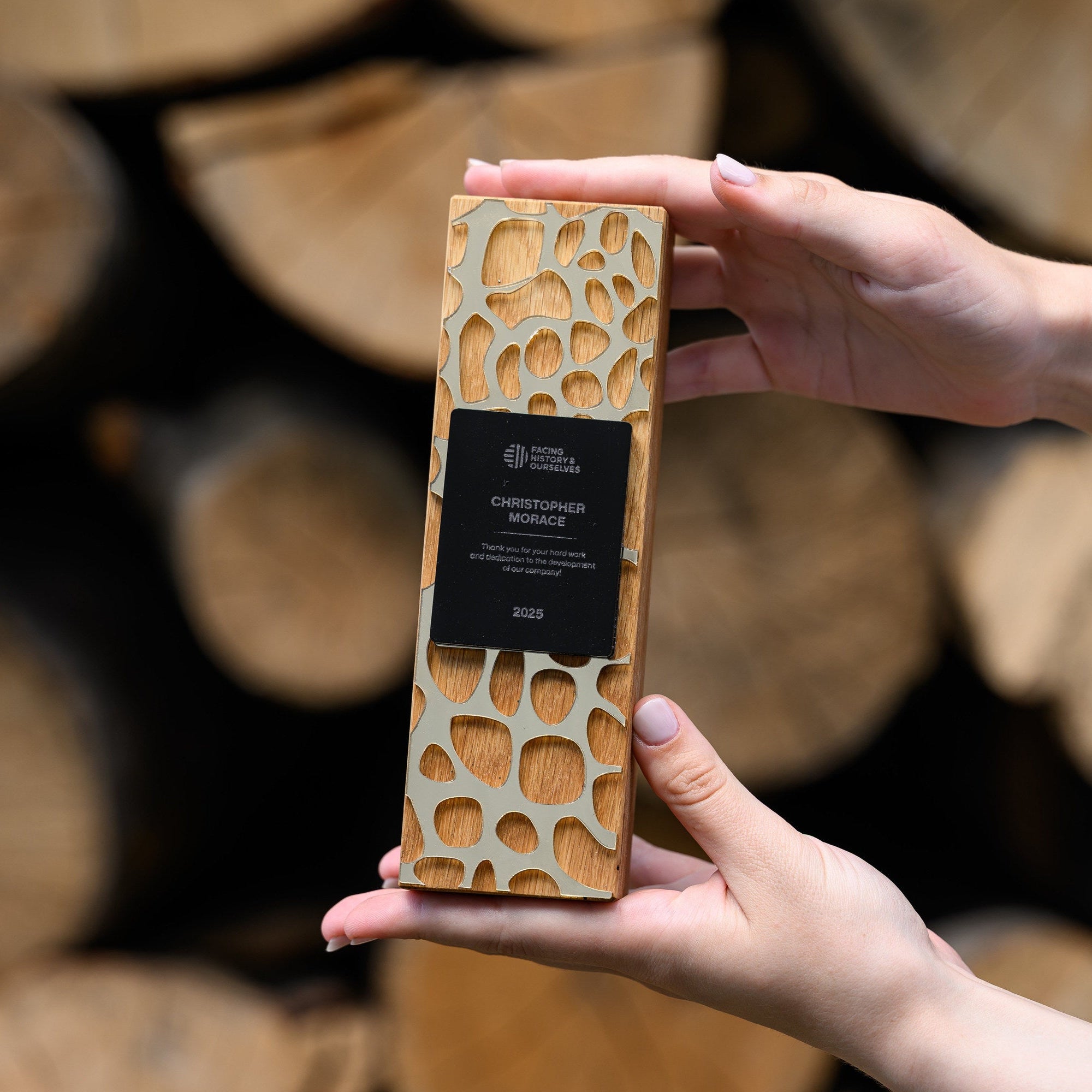 Hands holding an oak recognition award with gold acrylic lattice in front of stacked firewood logs