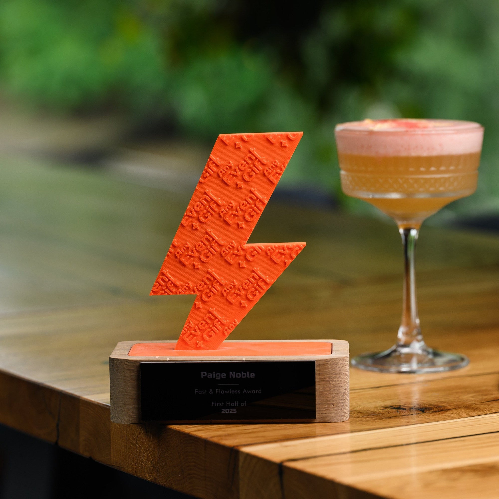 Orange 3D printed award standing on a bar counter beside a cocktail glass in a garden setting