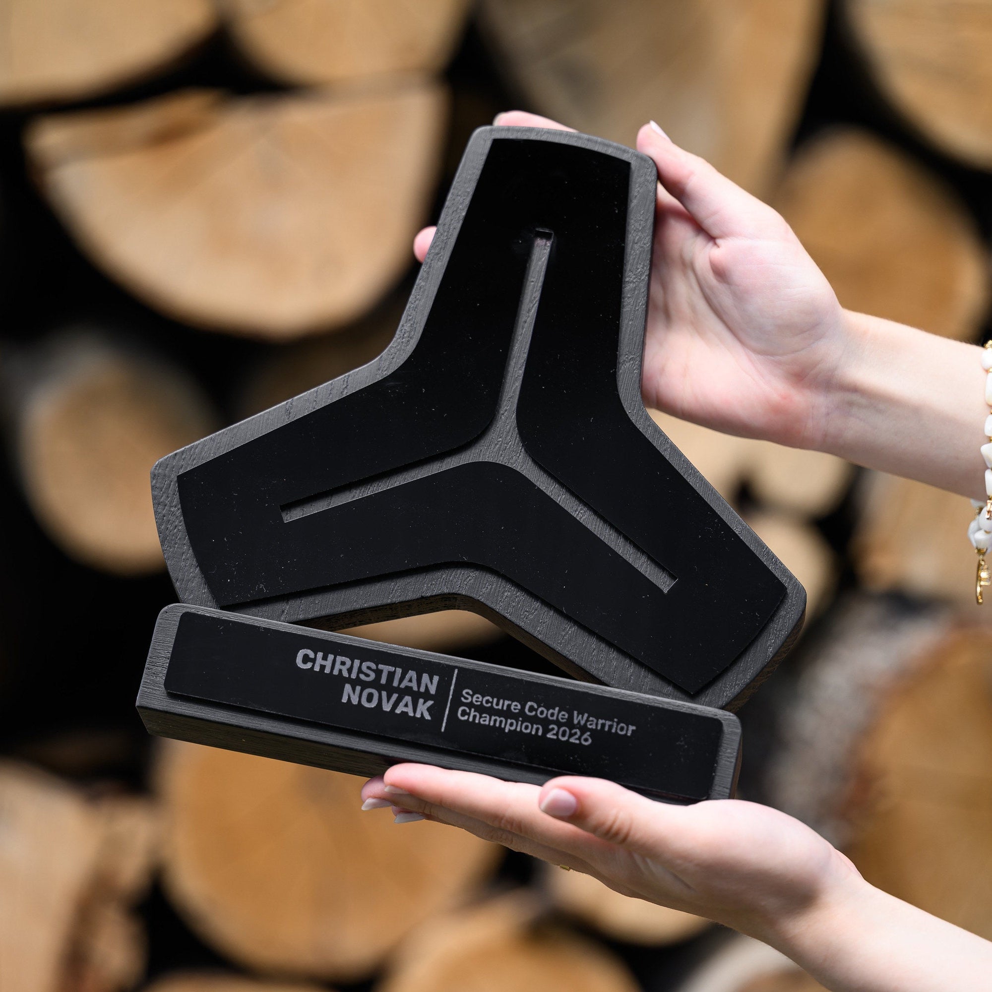 Hands holding a black oak and acrylic logo shaped award in front of a stack of cut firewood logs