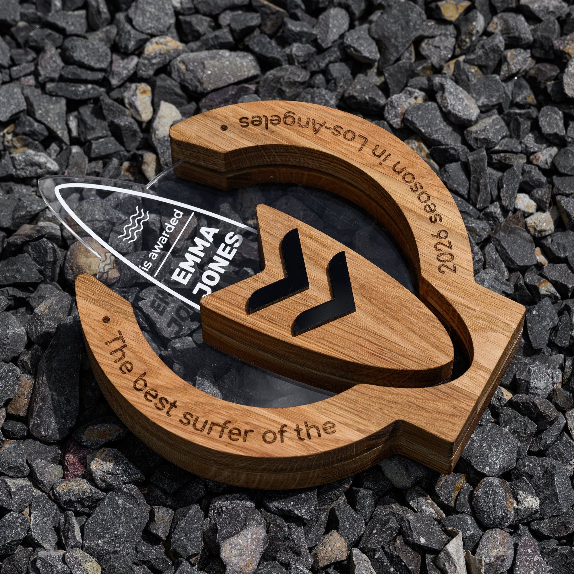 Angled view of the wooden surfing trophy on grey gravel showing sample engraving around the oak ring