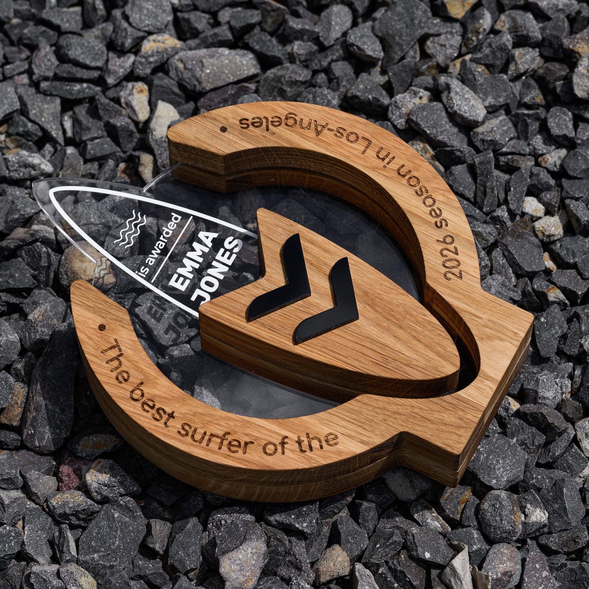 Oak surf award with printed acrylic surfboard lying face up on grey gravel, engraved ring visible
