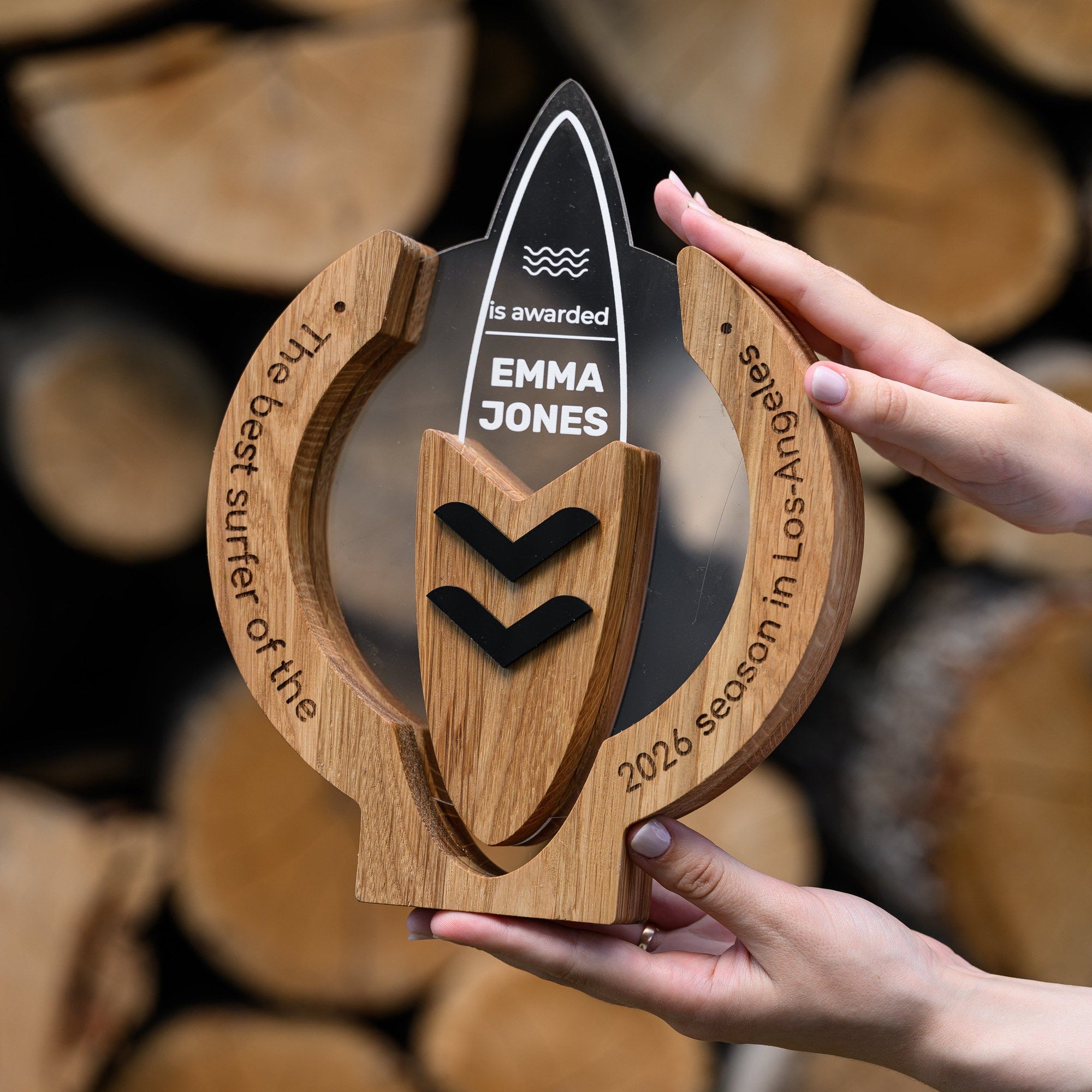 Hands holding an oak and acrylic surf trophy with black chevron inlays in front of stacked firewood