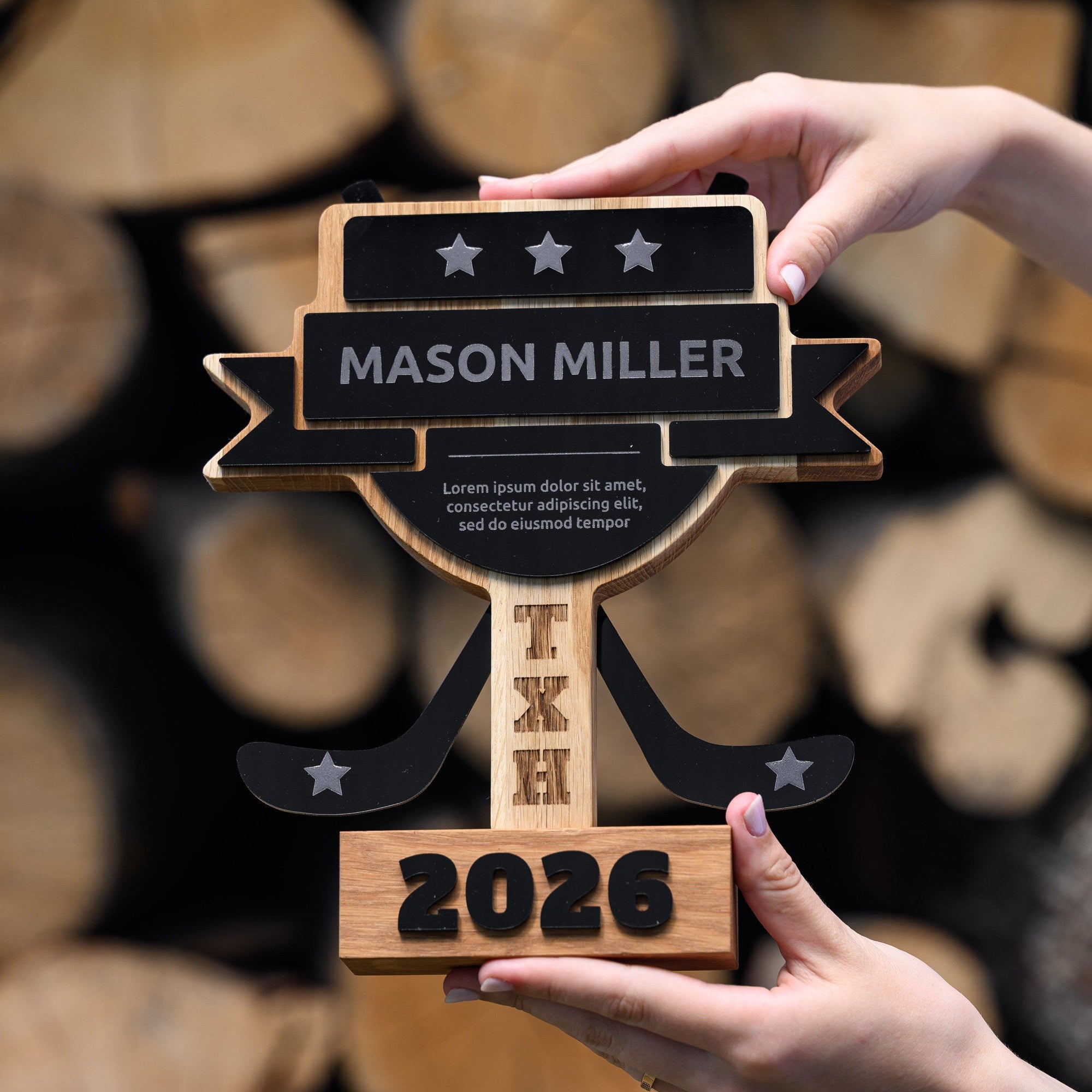 Hands holding an oak hockey trophy with engraved acrylic plates in front of stacked firewood logs
